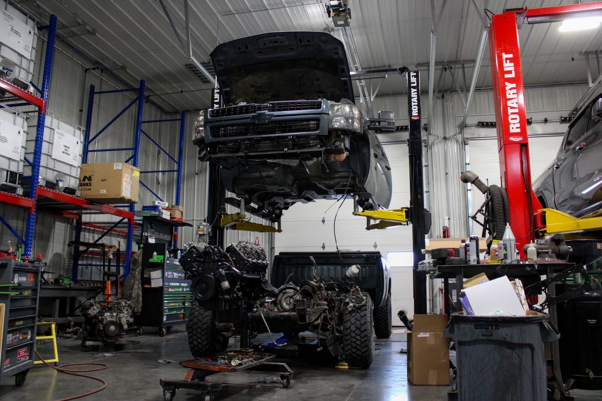 A vehicle chassis suspended on a lift in an auto repair shop, with an engine assembly on a stand below.