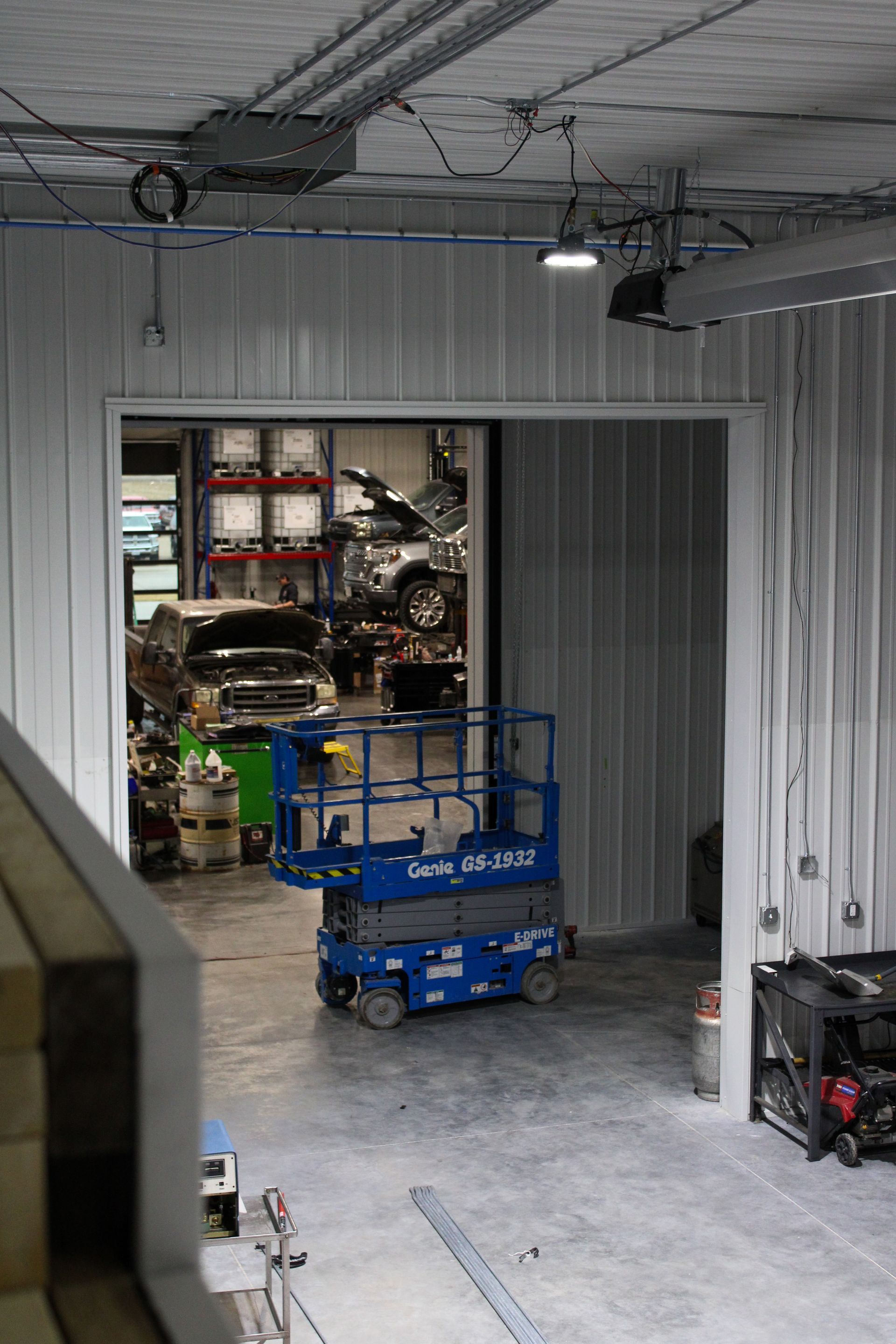A blue scissor lift parked in a warehouse workshop with cars visible in the background through a doorway.