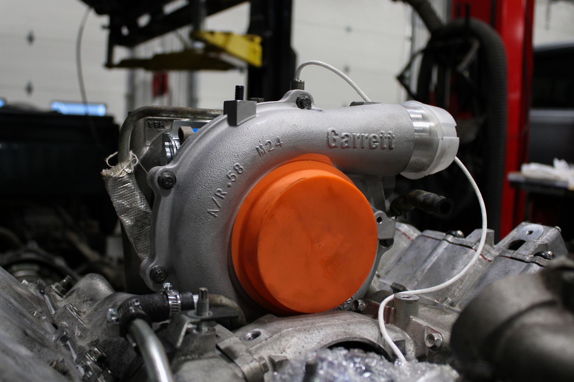 A silver Garrett turbocharger with a bright orange cap sits on an engine block inside a repair shop.