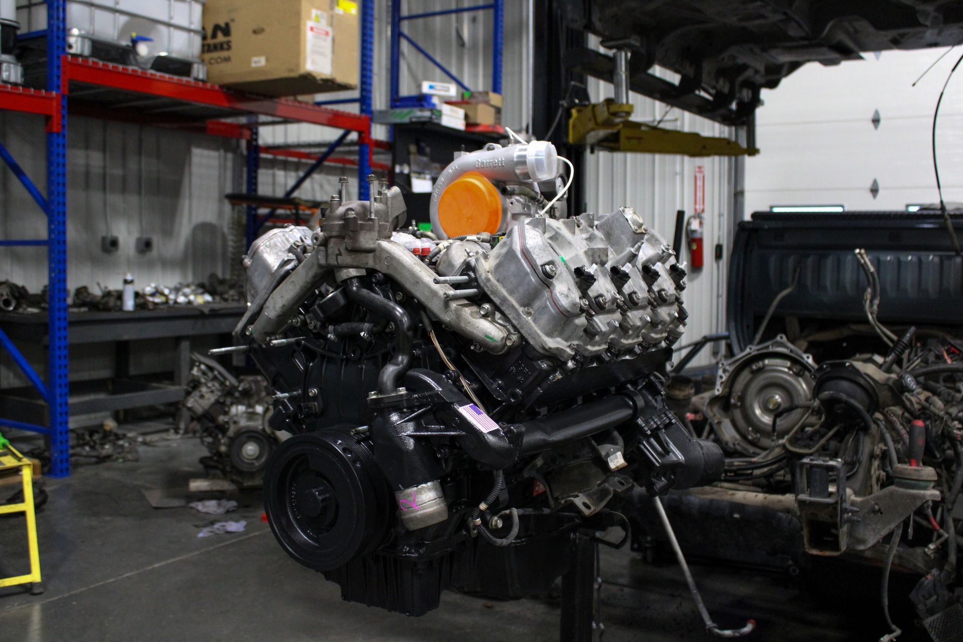 A large V8 automotive engine sitting on a stand in a workshop with metal storage shelving in the background.