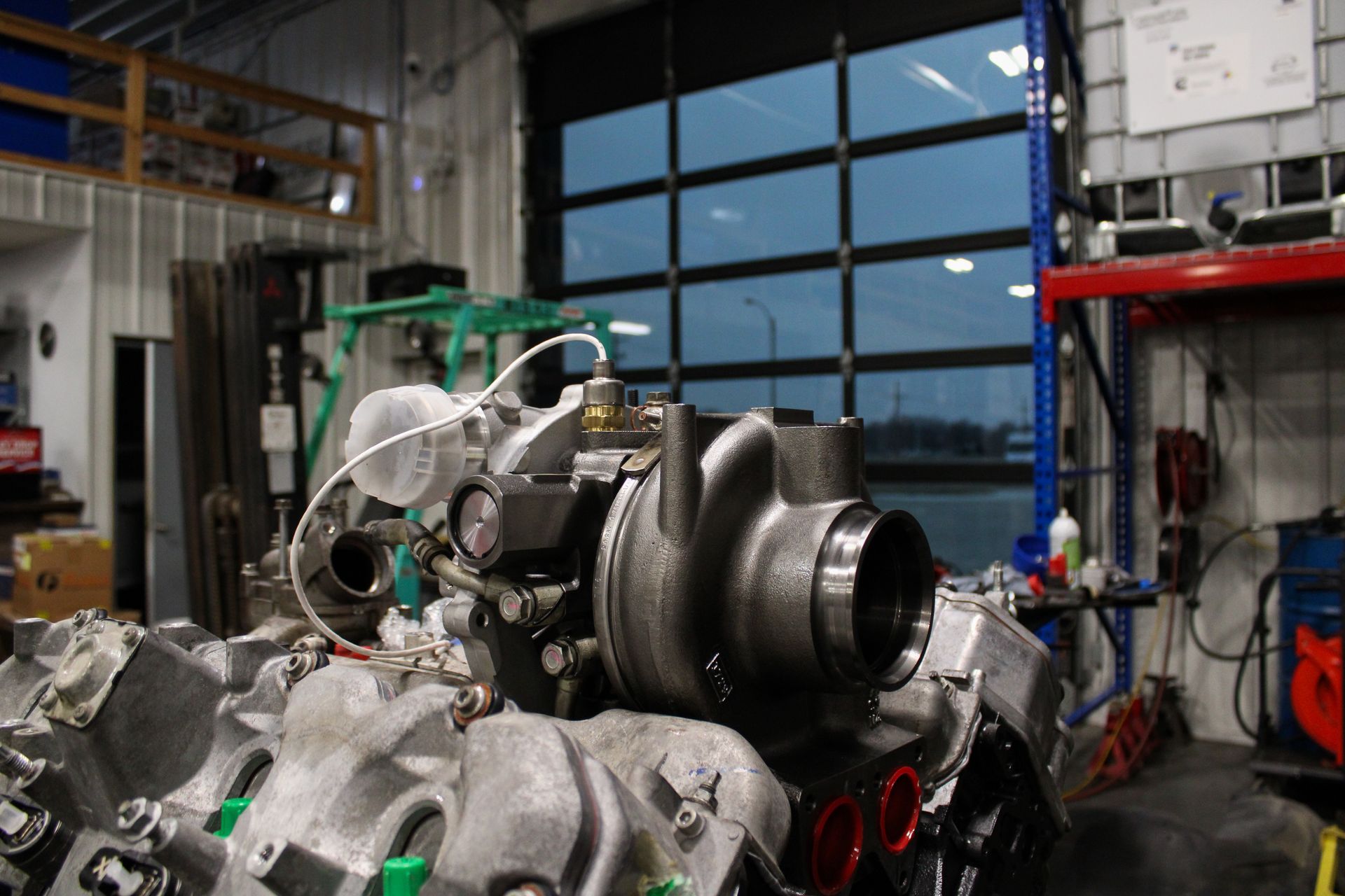 A close-up of a silver turbocharger mounted on an engine block inside a garage or mechanical workshop.
