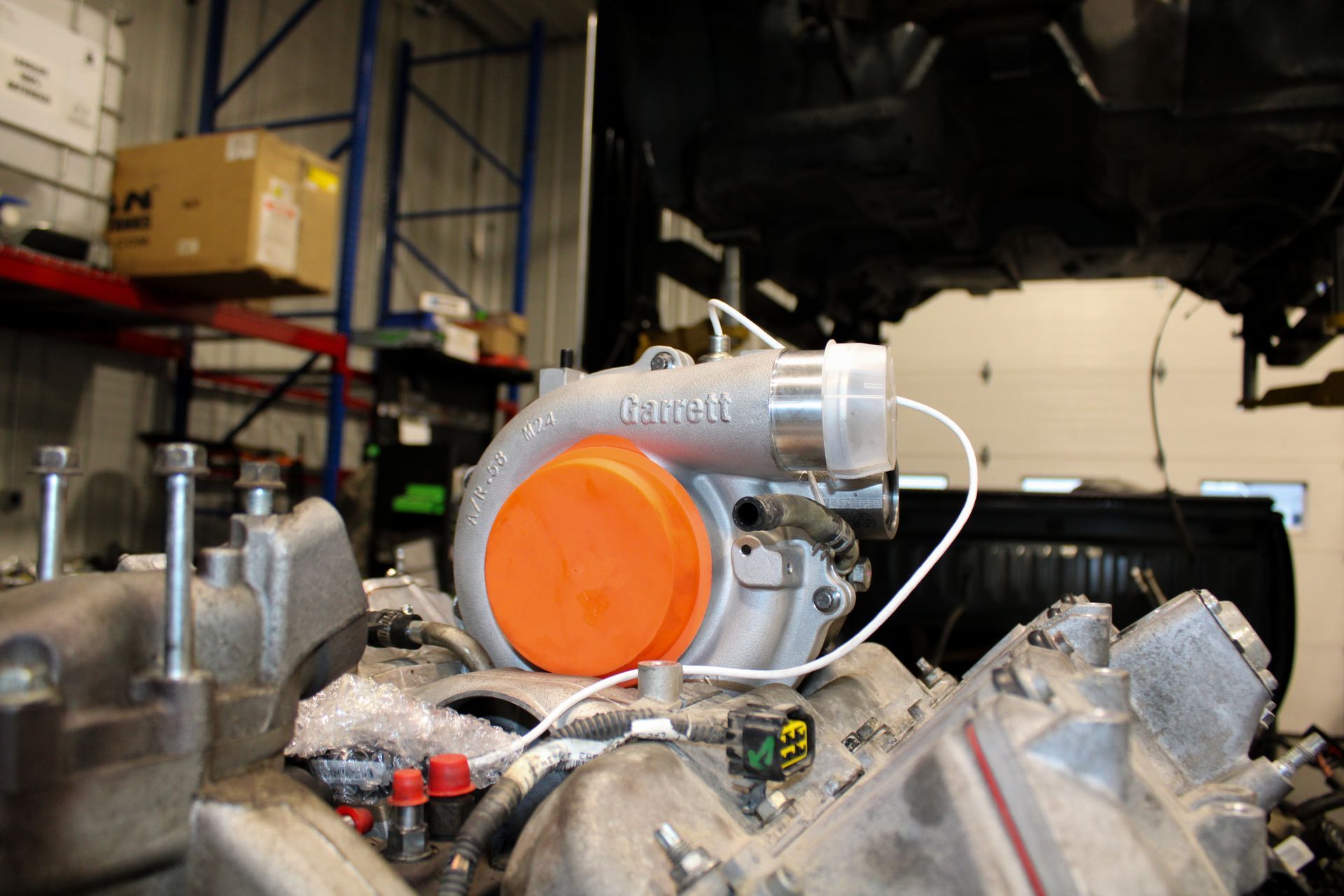 A metallic turbocharger with an orange cap sits on an engine block inside an automotive workshop.