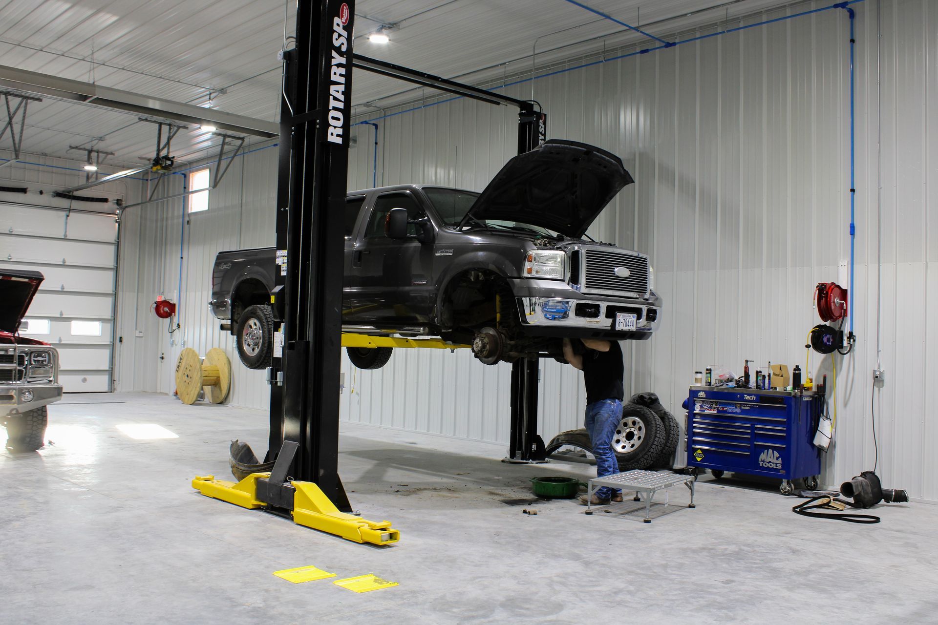 A mechanic works under a dark pickup truck raised on a two-post lift inside an auto repair shop.