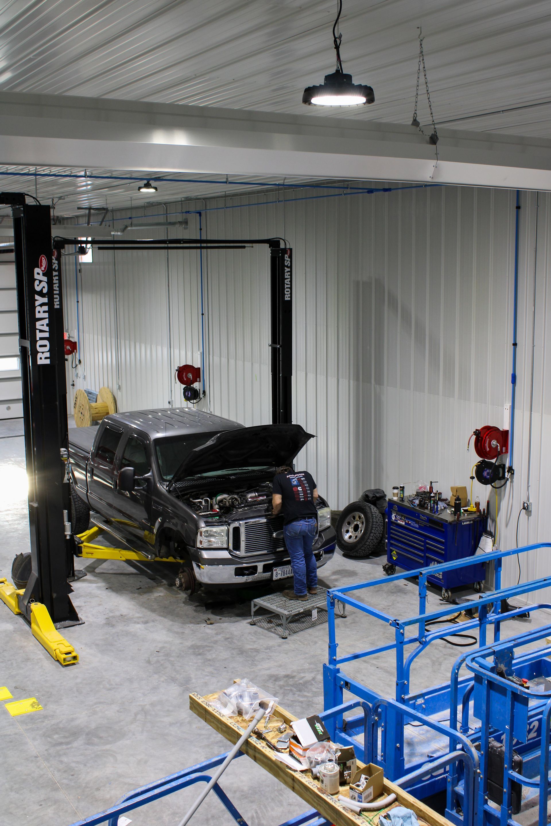 A mechanic works under the hood of a black pickup truck raised on a hydraulic lift in a bright industrial garage.