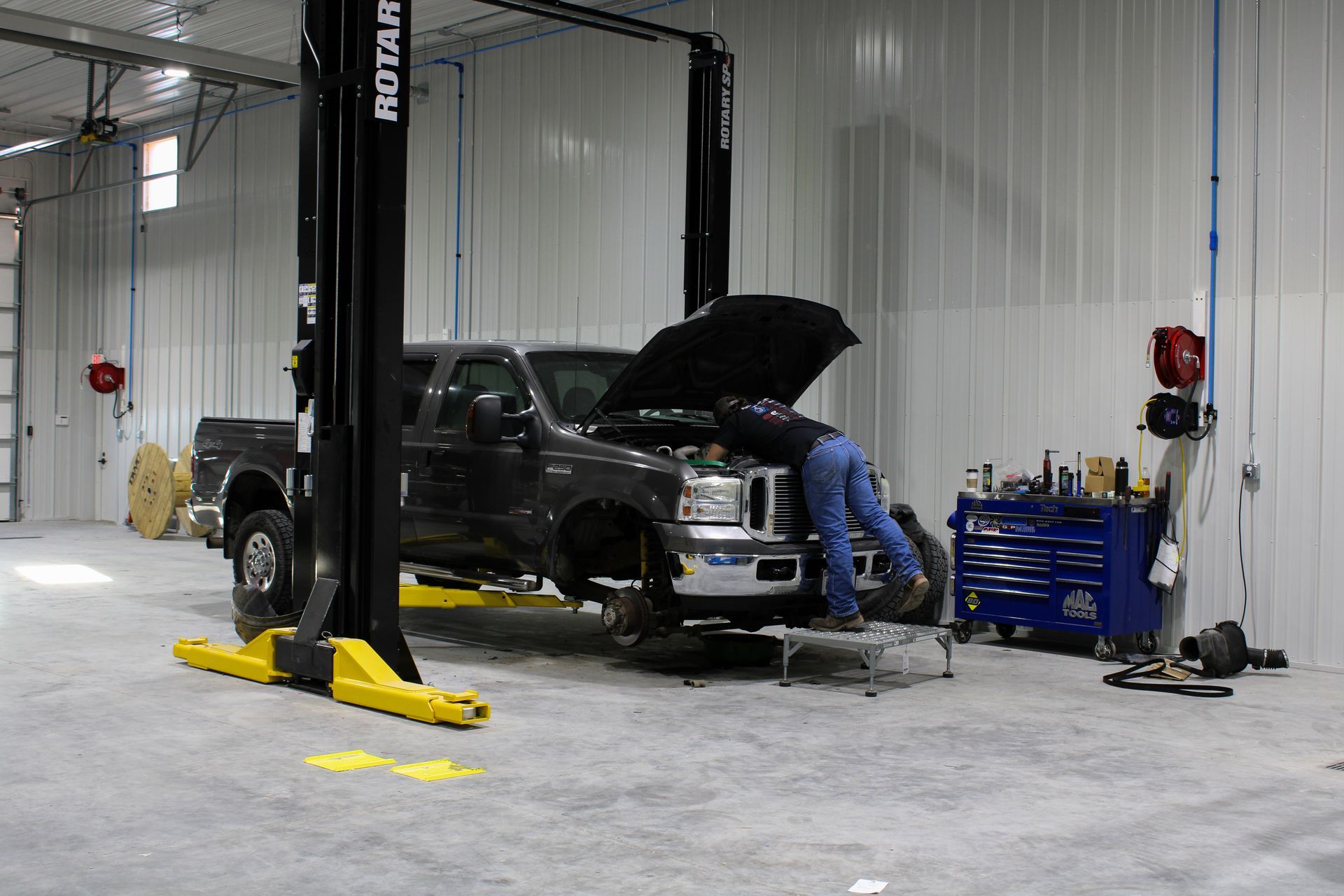 A mechanic works on the engine of a dark pickup truck raised on a hydraulic lift in a bright workshop.
