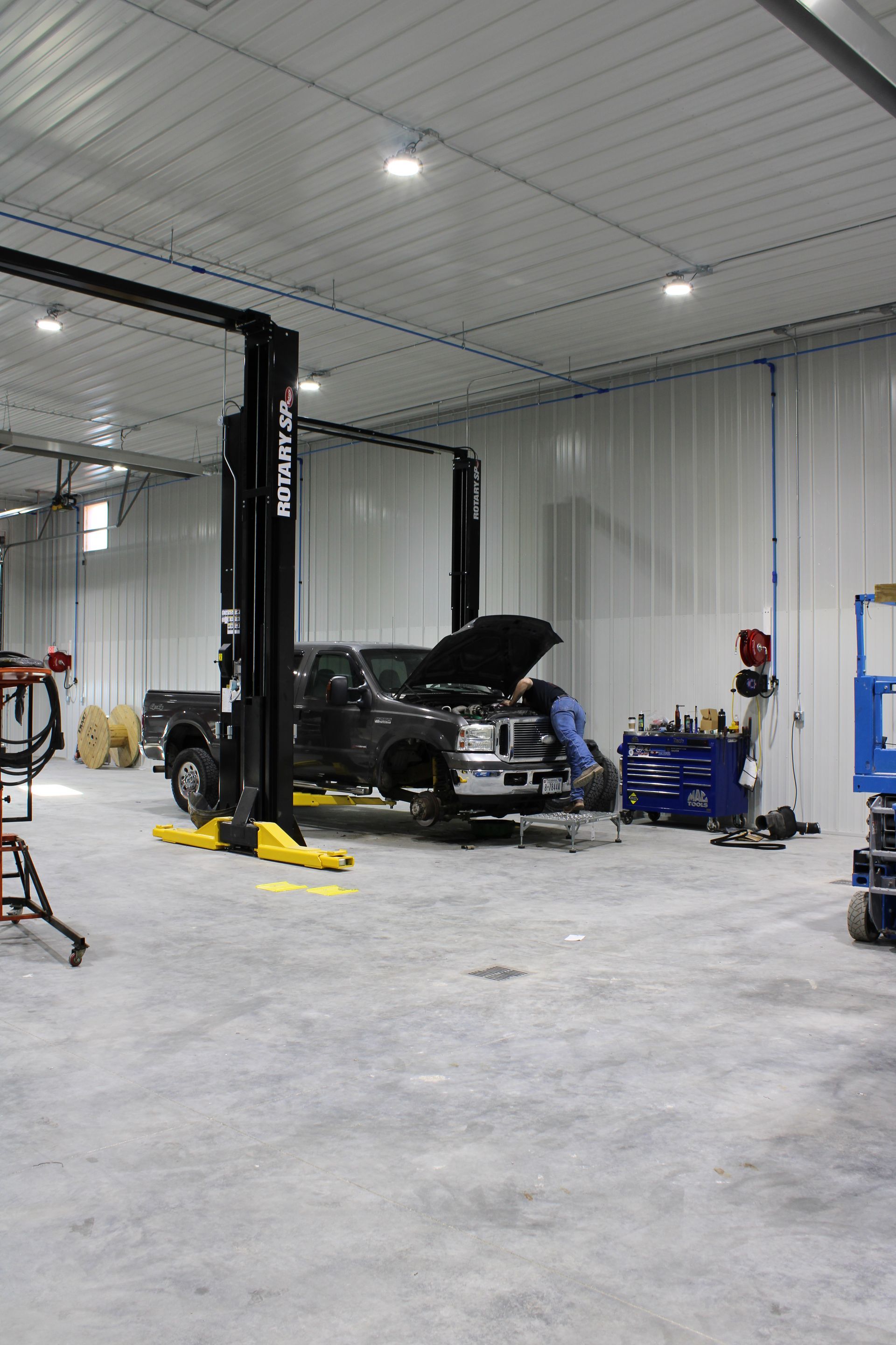 A mechanic works on a truck inside a brightly lit auto shop with a tall vehicle lift and blue tool chest.