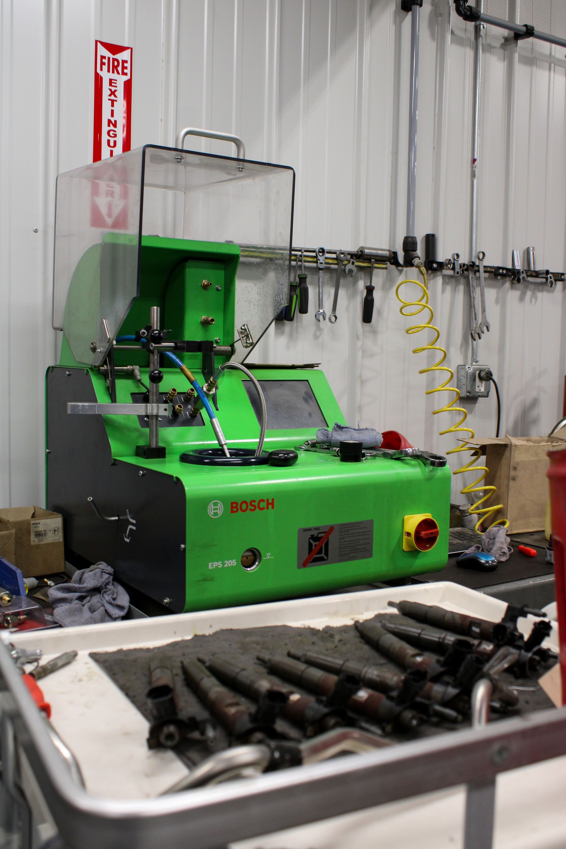 A green Bosch diagnostic machine sits on a workbench with automotive fuel injectors arranged in a tray in the foreground.
