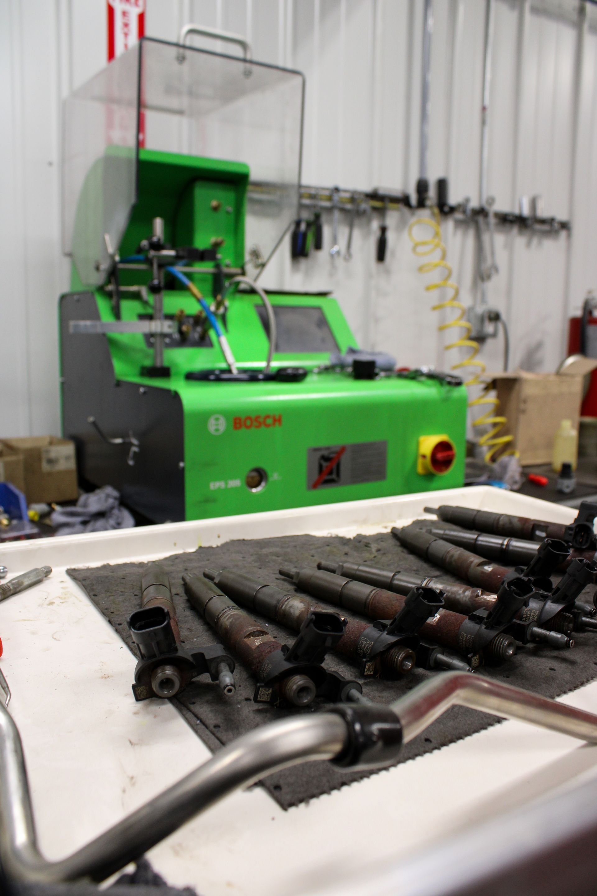 A row of automotive fuel injectors sits on a tray in front of a green Bosch diesel fuel injector test bench.