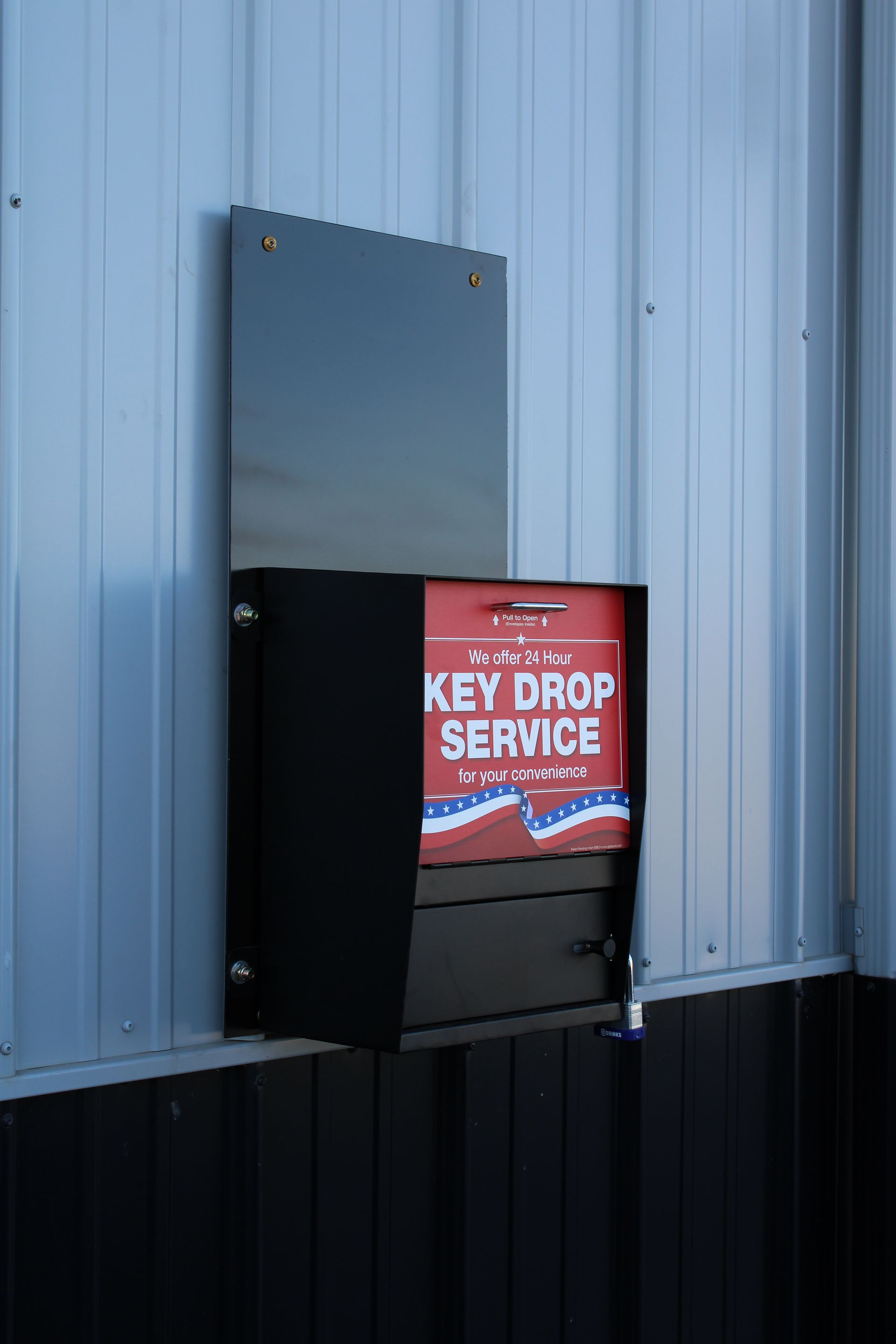 A black metal key drop service box mounted on a blue corrugated metal wall with red and white signage.