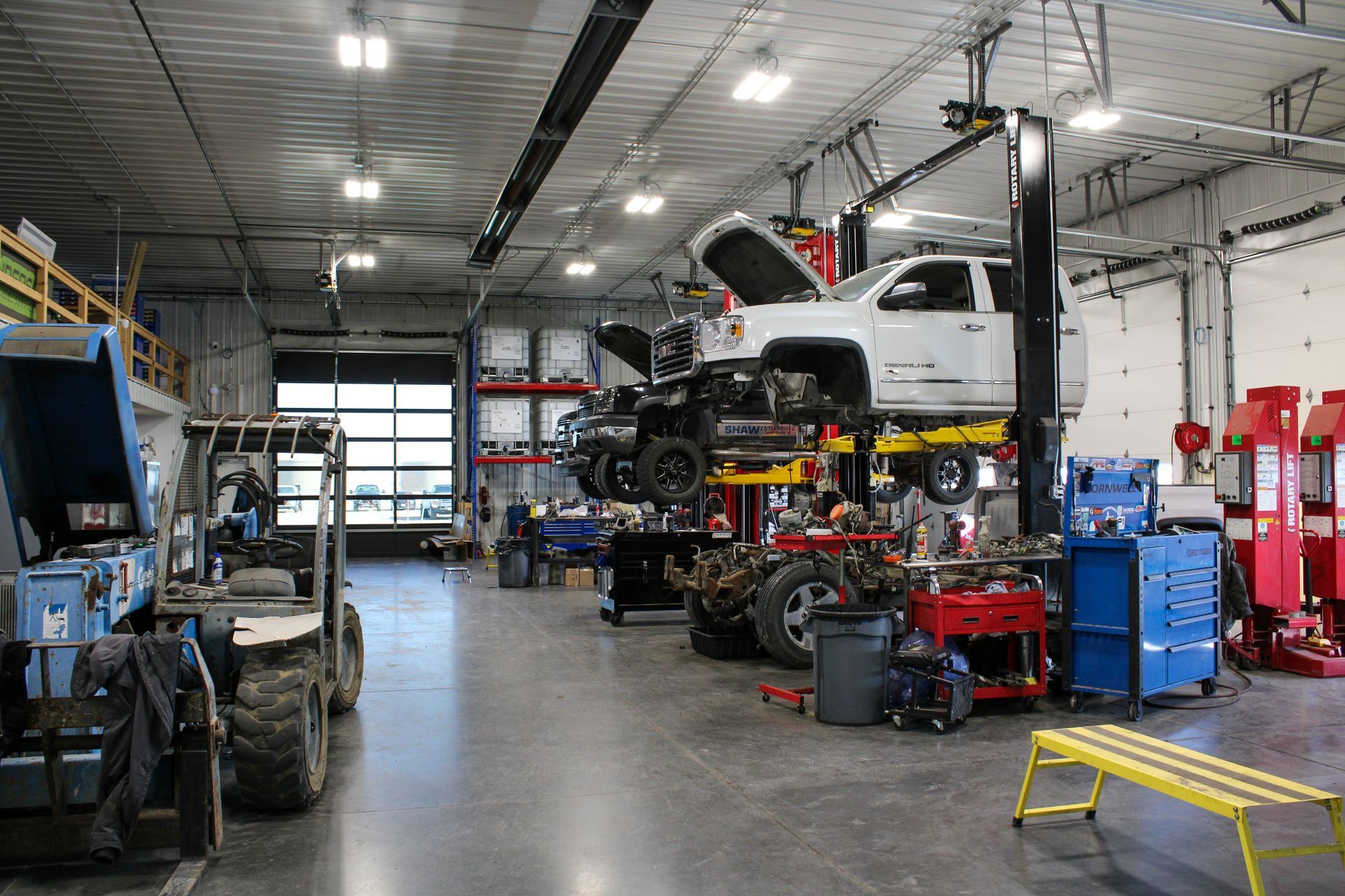 A brightly lit automotive repair shop with vehicles lifted for maintenance, work benches, and equipment on a concrete floor.
