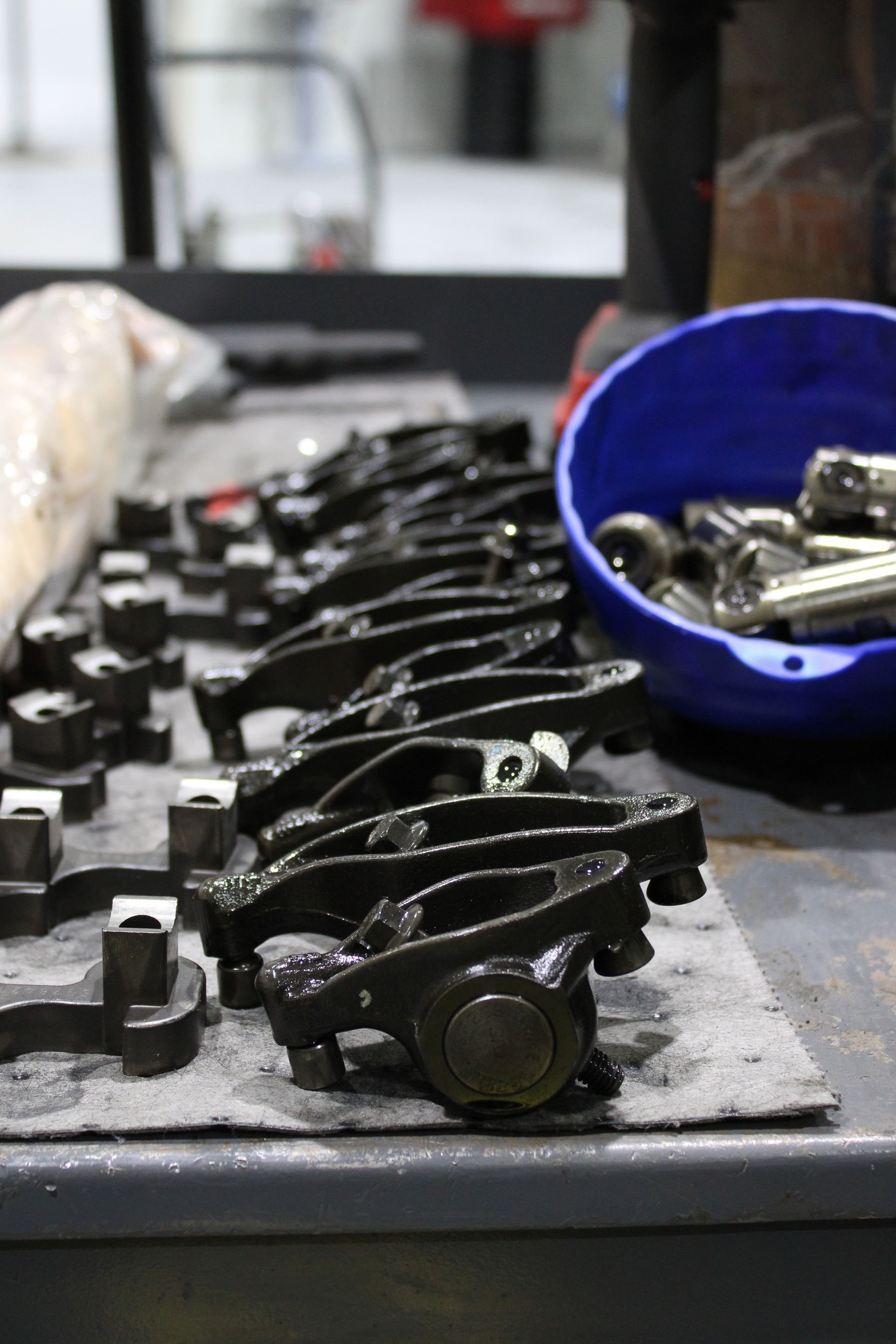 A row of metal engine rocker arms arranged on a workbench next to a blue bowl containing small mechanical parts.