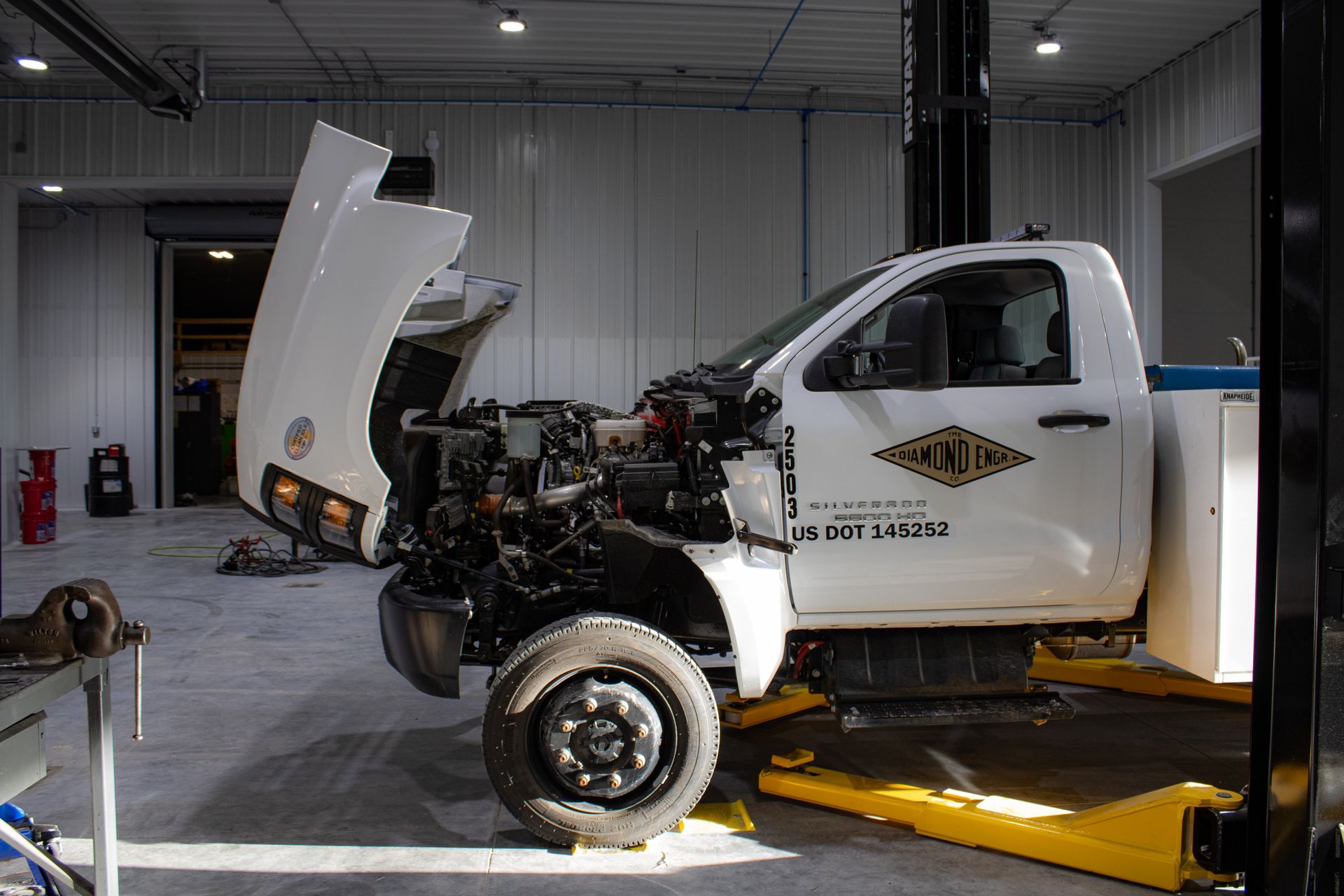 A white service truck with its hood open, positioned on a lift inside a well-lit auto repair shop.