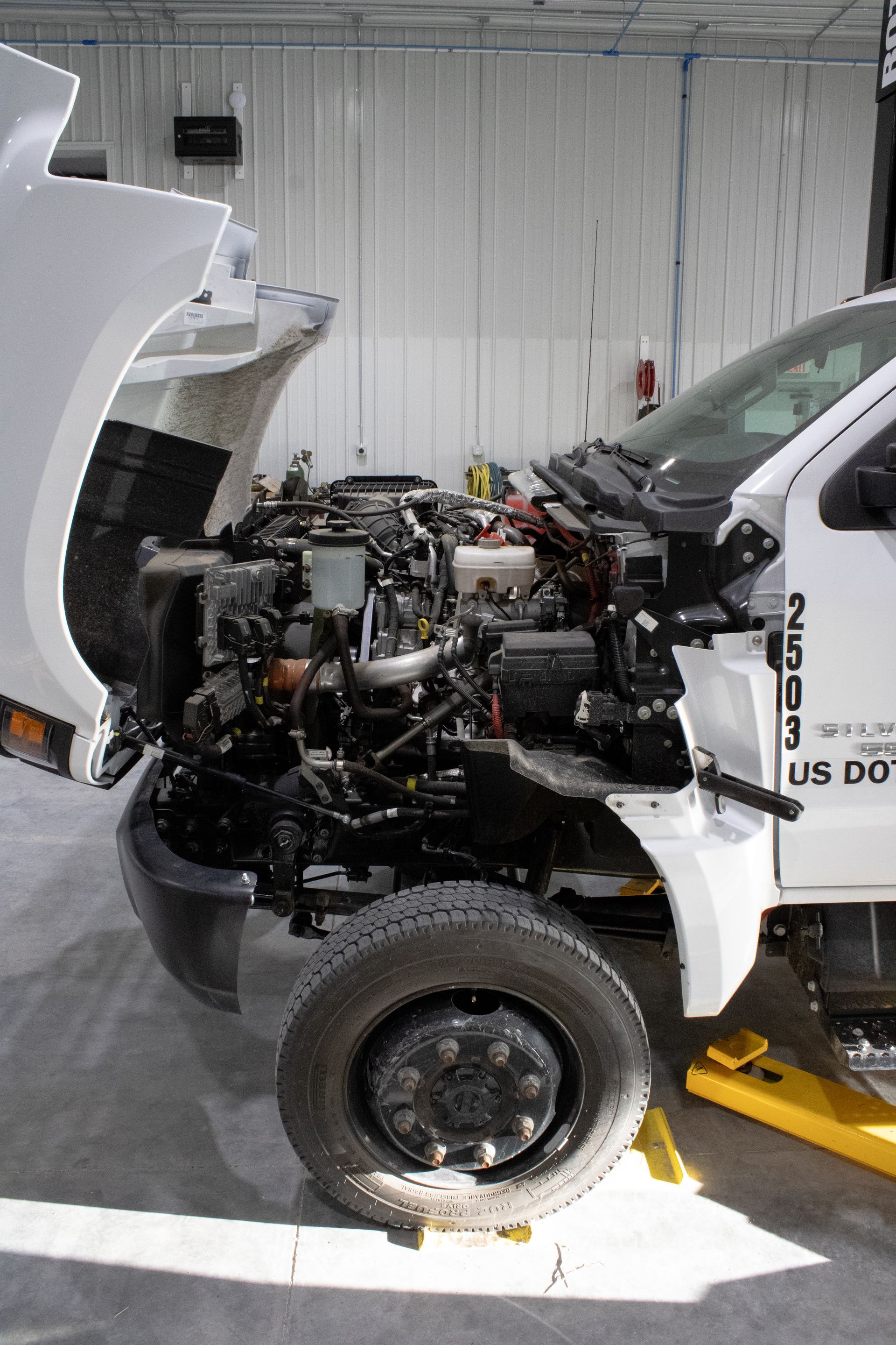 A white commercial truck parked inside a repair shop with its hood open, exposing the engine bay for maintenance.
