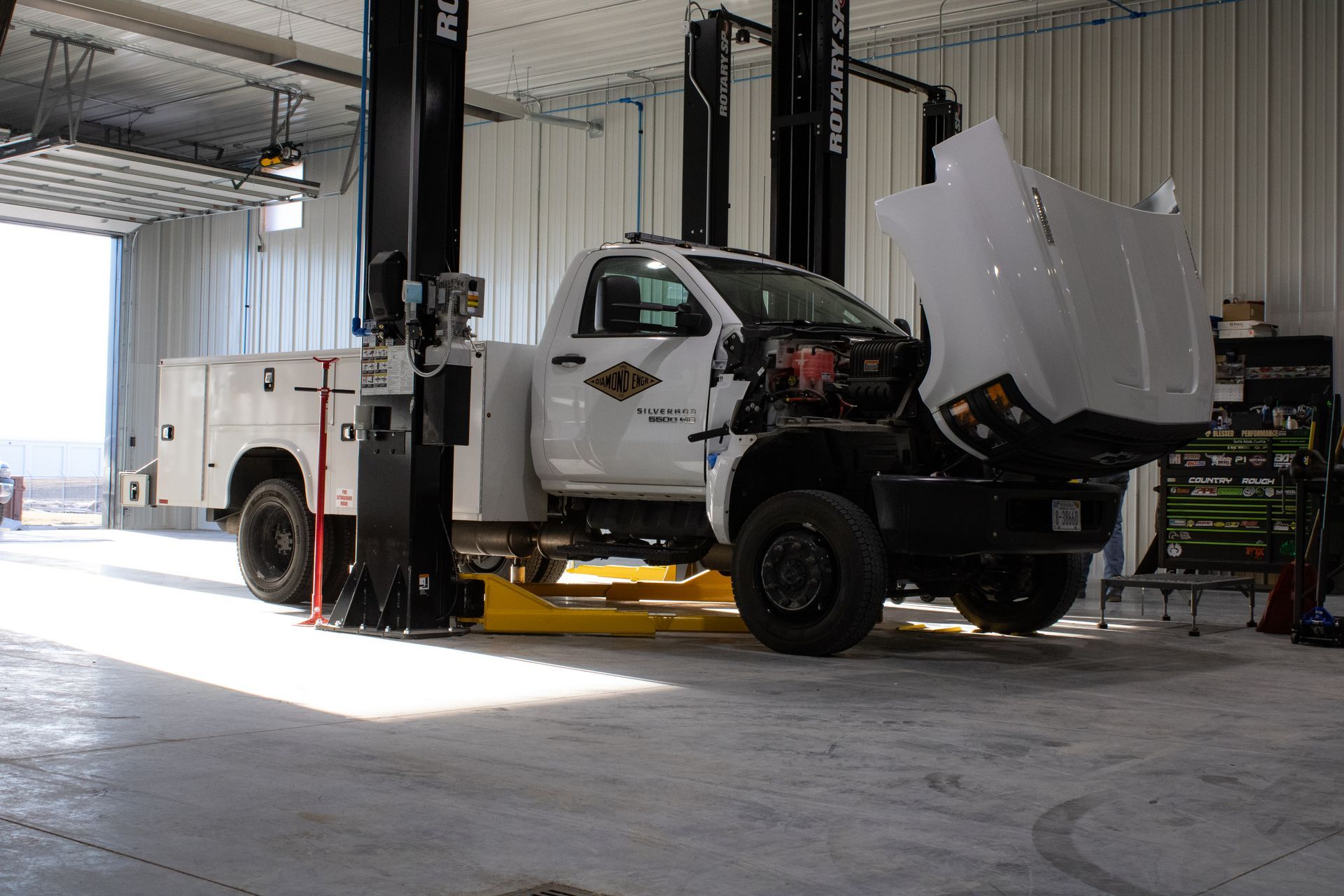 A white utility truck with its hood open is elevated on a lift inside a brightly lit vehicle maintenance garage.