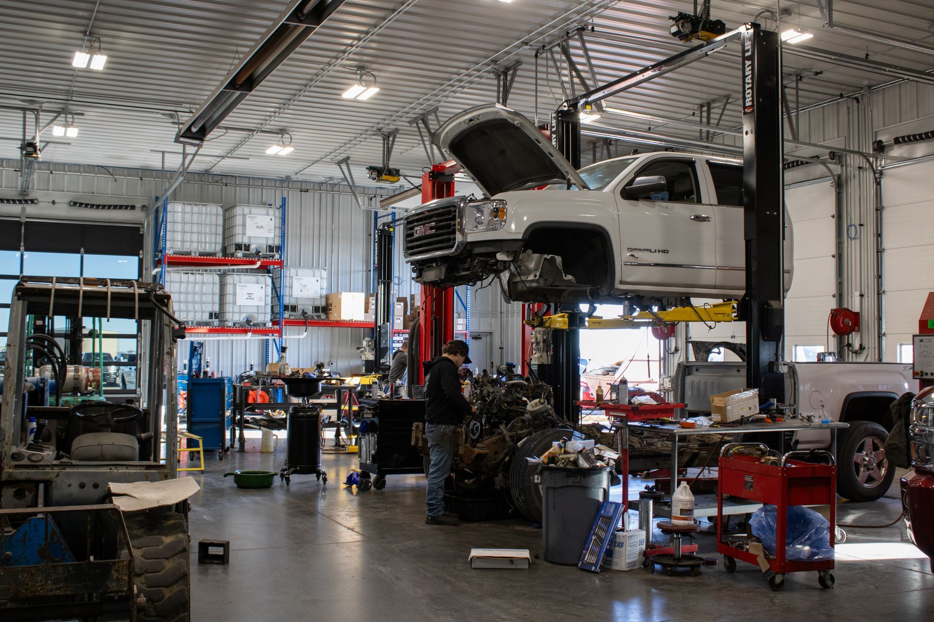 A mechanic works on a white truck lifted on a hydraulic rack inside a bright, industrial automotive repair shop.
