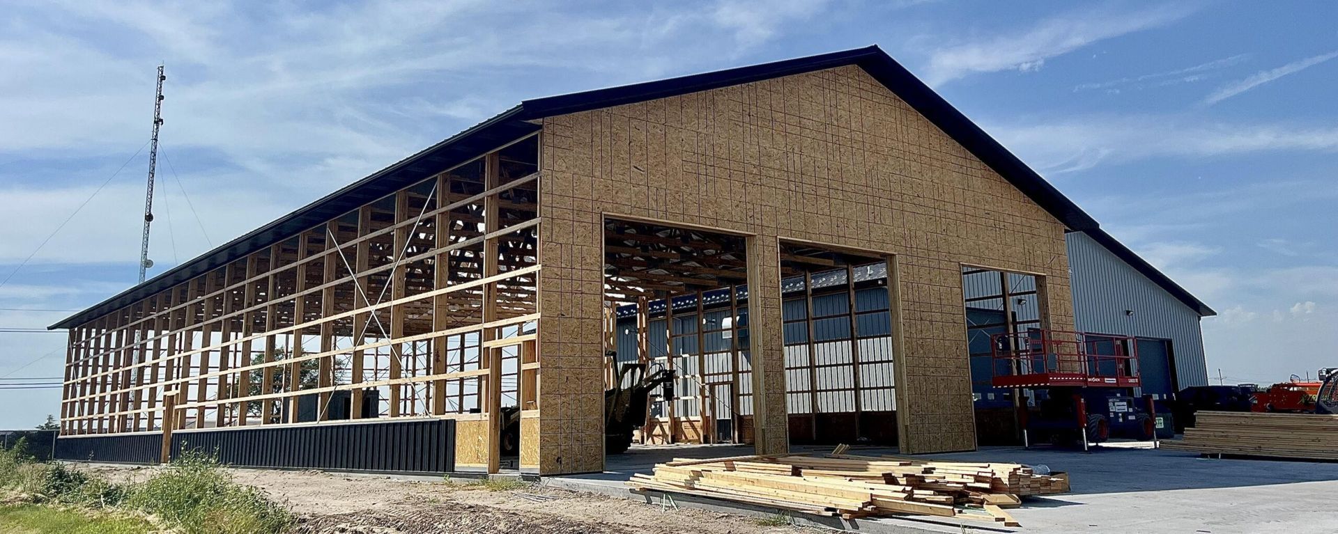 A partially constructed, long wooden-framed building with exposed wall studs and roof trusses under a bright blue sky.
