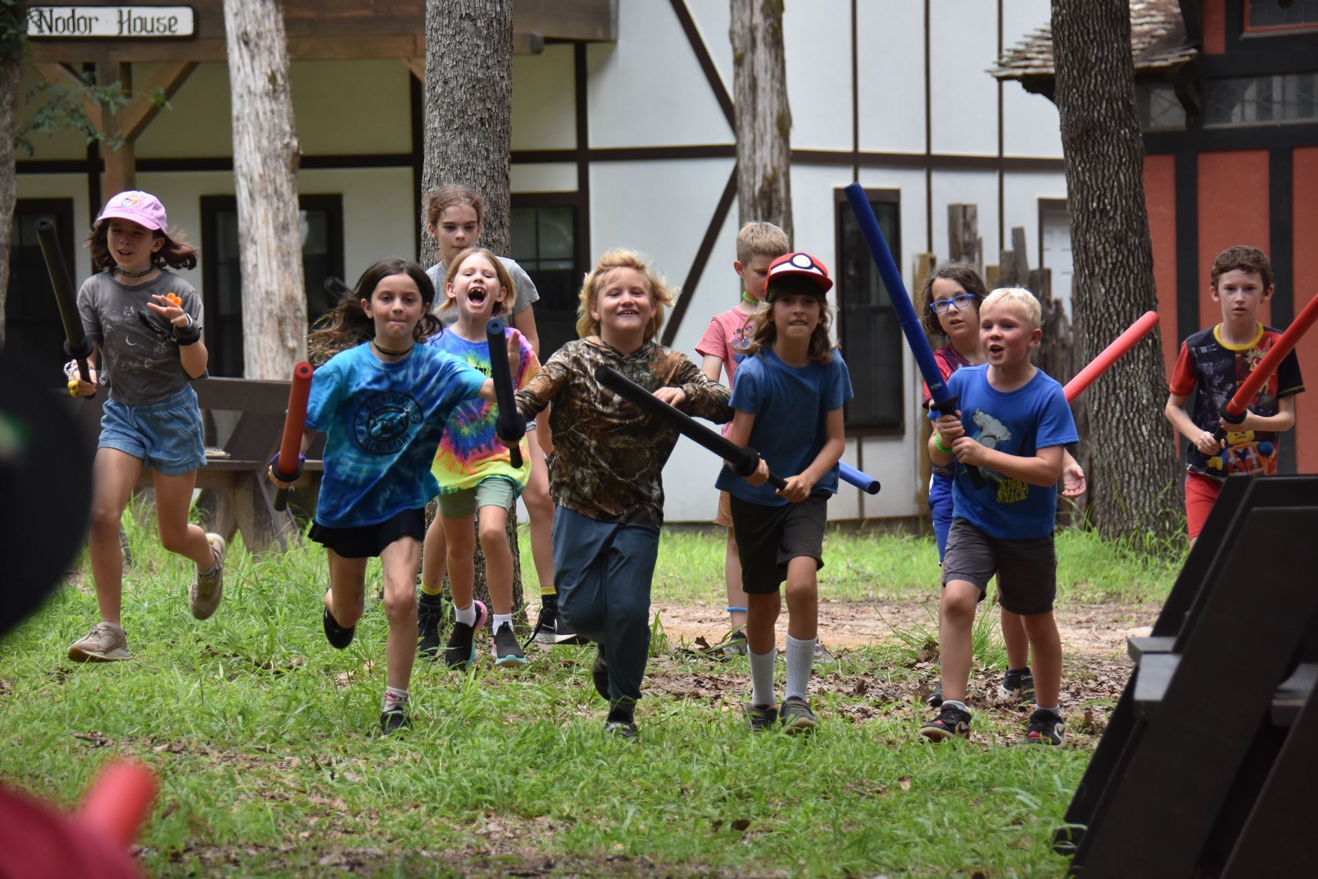 Kids in a grassy area with lightsabers, running toward the camera, with a building in the background.
