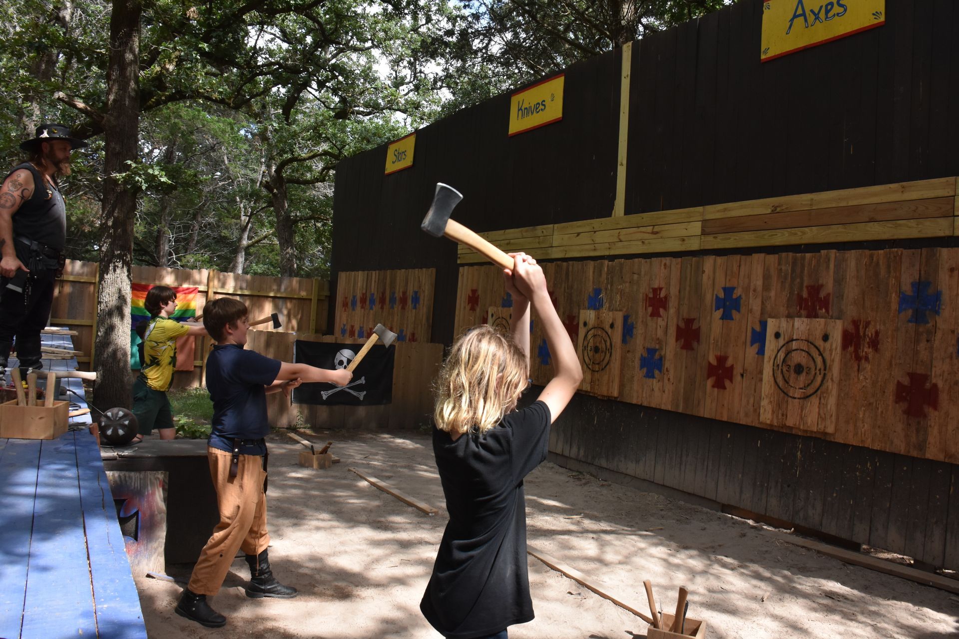 Two children throwing axes at wooden targets at an outdoor axe-throwing range; an instructor looks on.