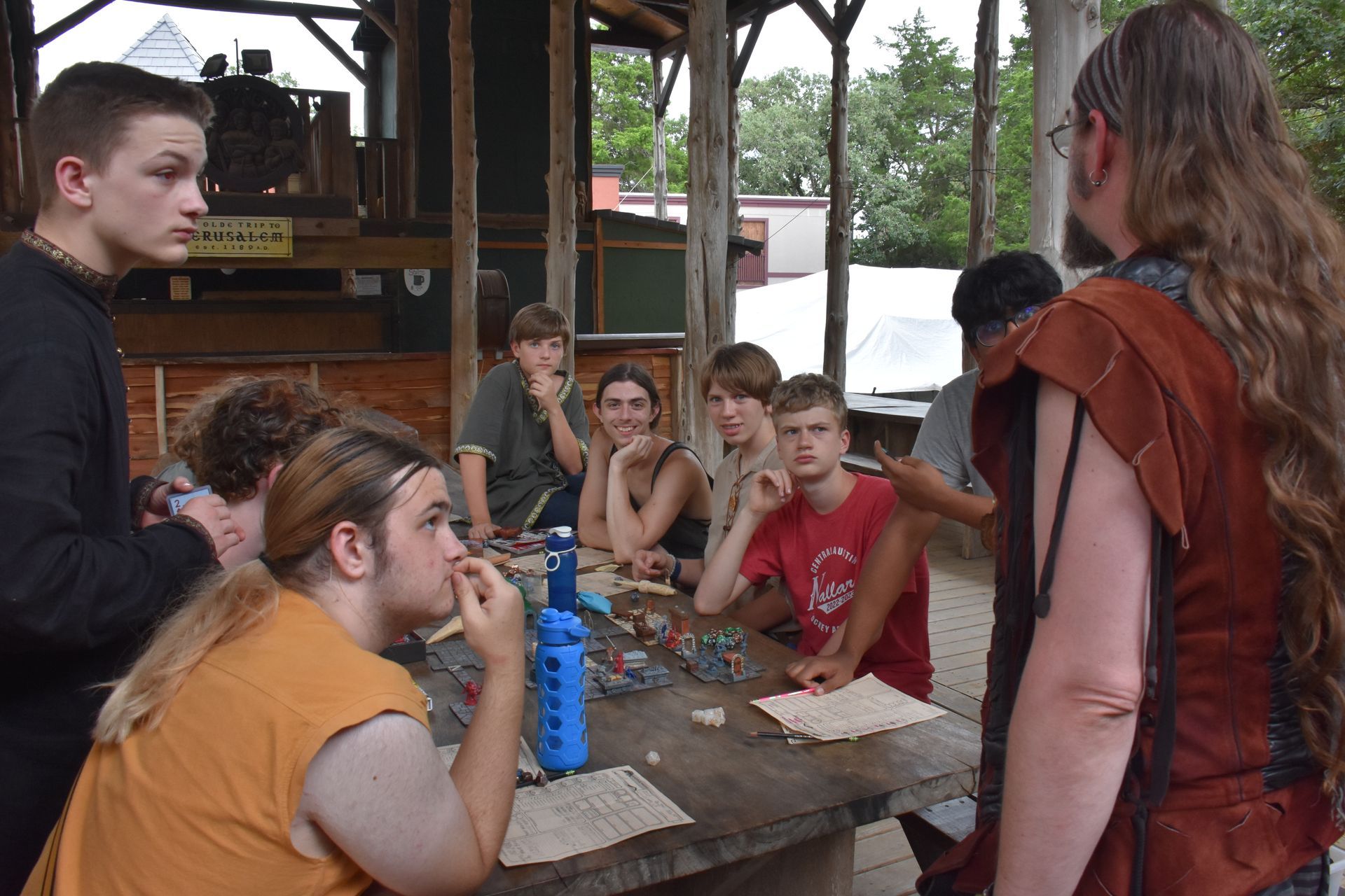 A group of people sit around a table, some looking at papers and others listening, outdoors under a wooden shelter.