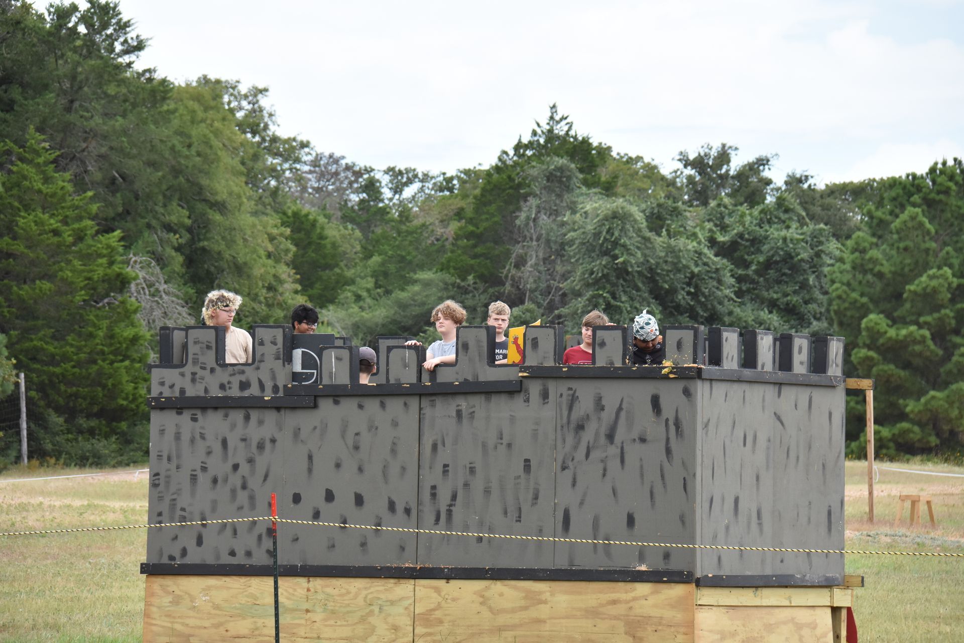 People in a gray castle-like structure in an outdoor field with green trees and sky in the background.