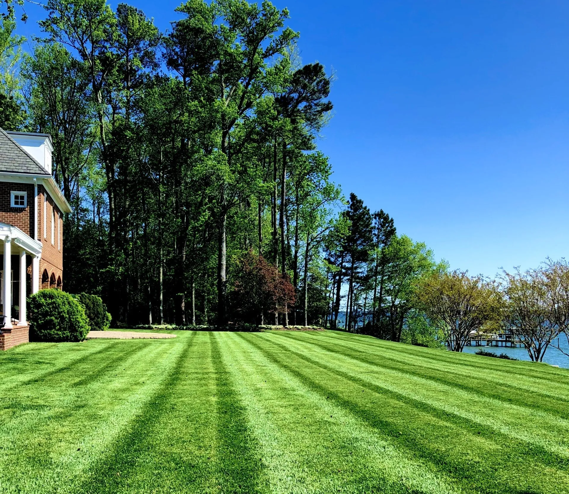 Well-manicured lawn with striped mowing pattern, trees, and partial view of a brick house and waterfront under a blue sky.