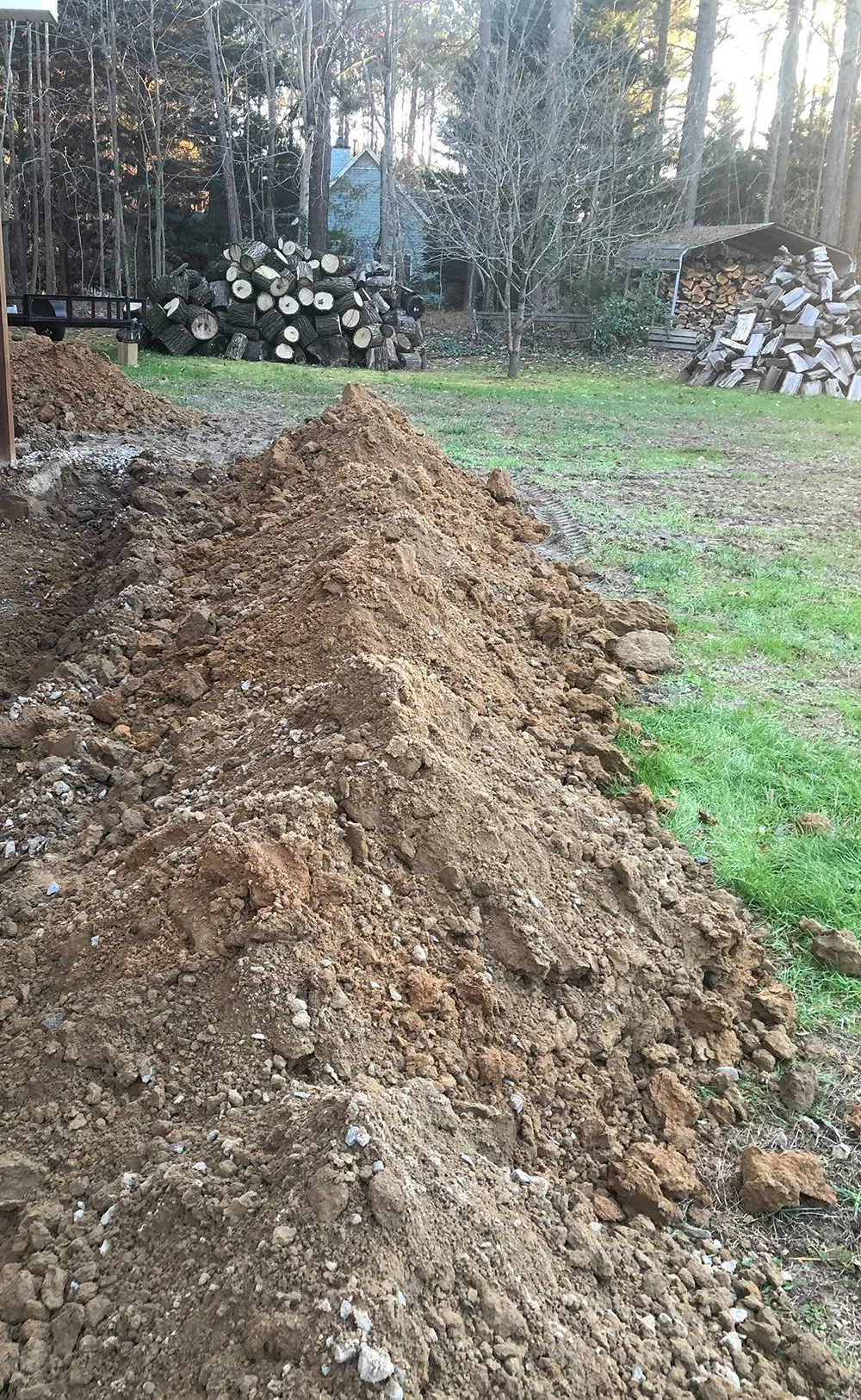 Pile of dirt along a yard's edge, possibly from digging; logs and trees in the background.