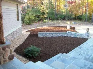 Exterior view of a house with stone steps, mulch, and a retaining wall in a yard, with trees in the background.