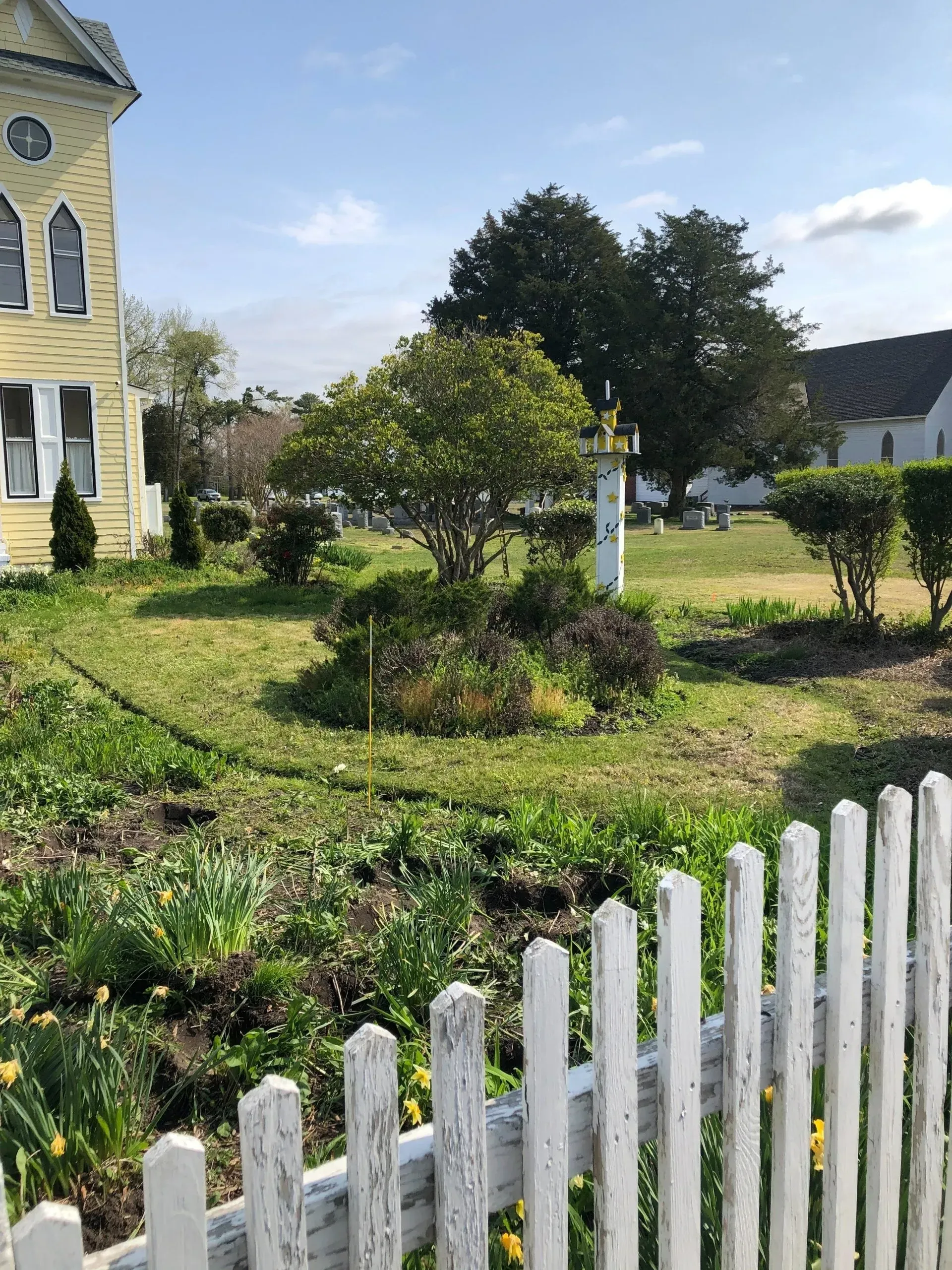 Yellow house with white picket fence, green yard, and small trees on a sunny day.