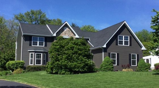 Two-story house with dark gray siding, multiple windows, and a green lawn under a blue sky.