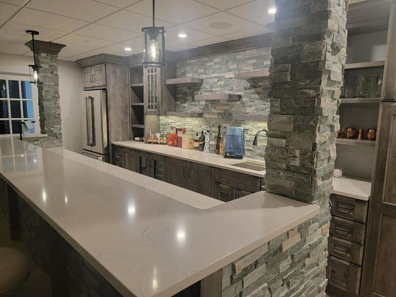 Basement bar with stone accents, wood cabinetry, and a countertop.  Lighting fixture hangs overhead.