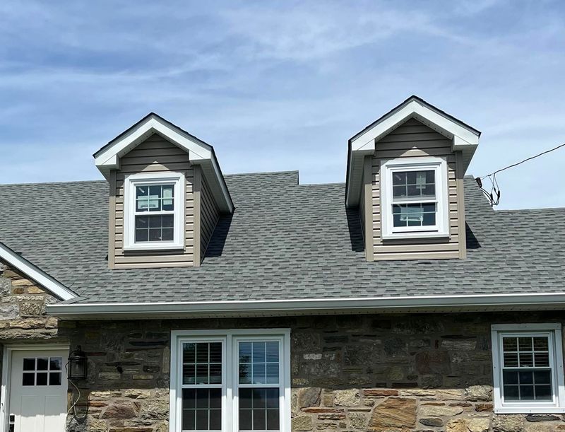 House with two dormer windows, stone facade, gray roof, and blue sky.