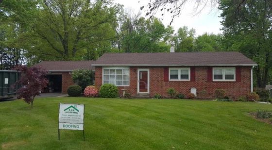 Red brick ranch house with a green lawn and brown roof. Roofing sign in front.