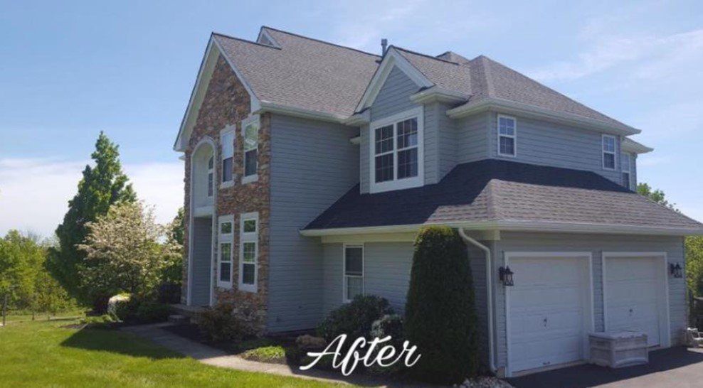 Two-story house with gray siding, stone facade, dark roof, and two-car garage under a bright blue sky.