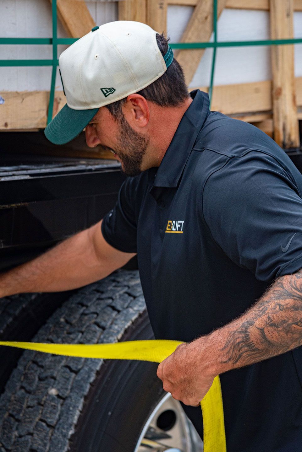 A man secures cargo with a yellow strap on a truck tire. He wears a hat and a black shirt outdoors.