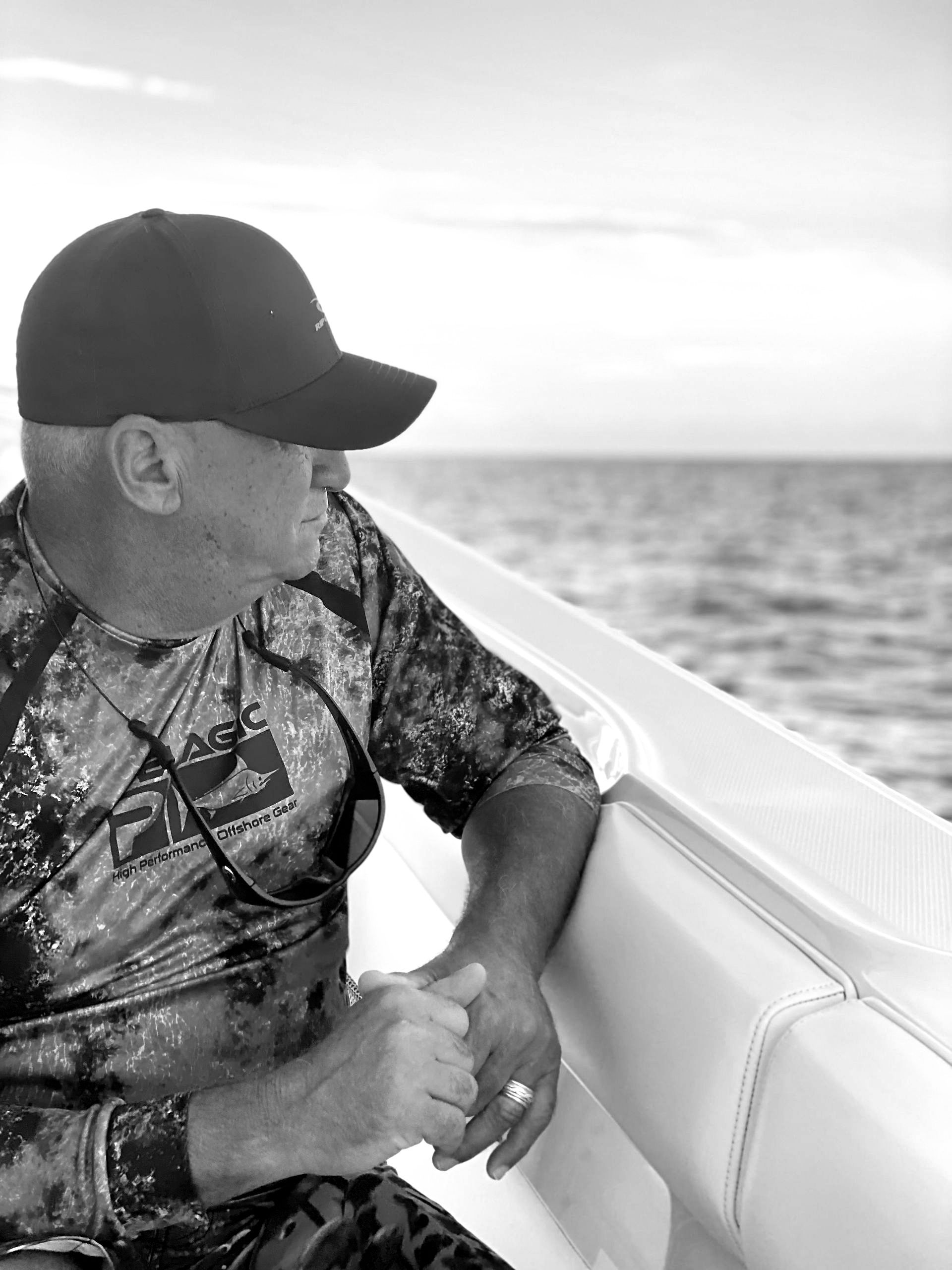 Man on boat, looking out at sea, wearing hat and camo shirt.