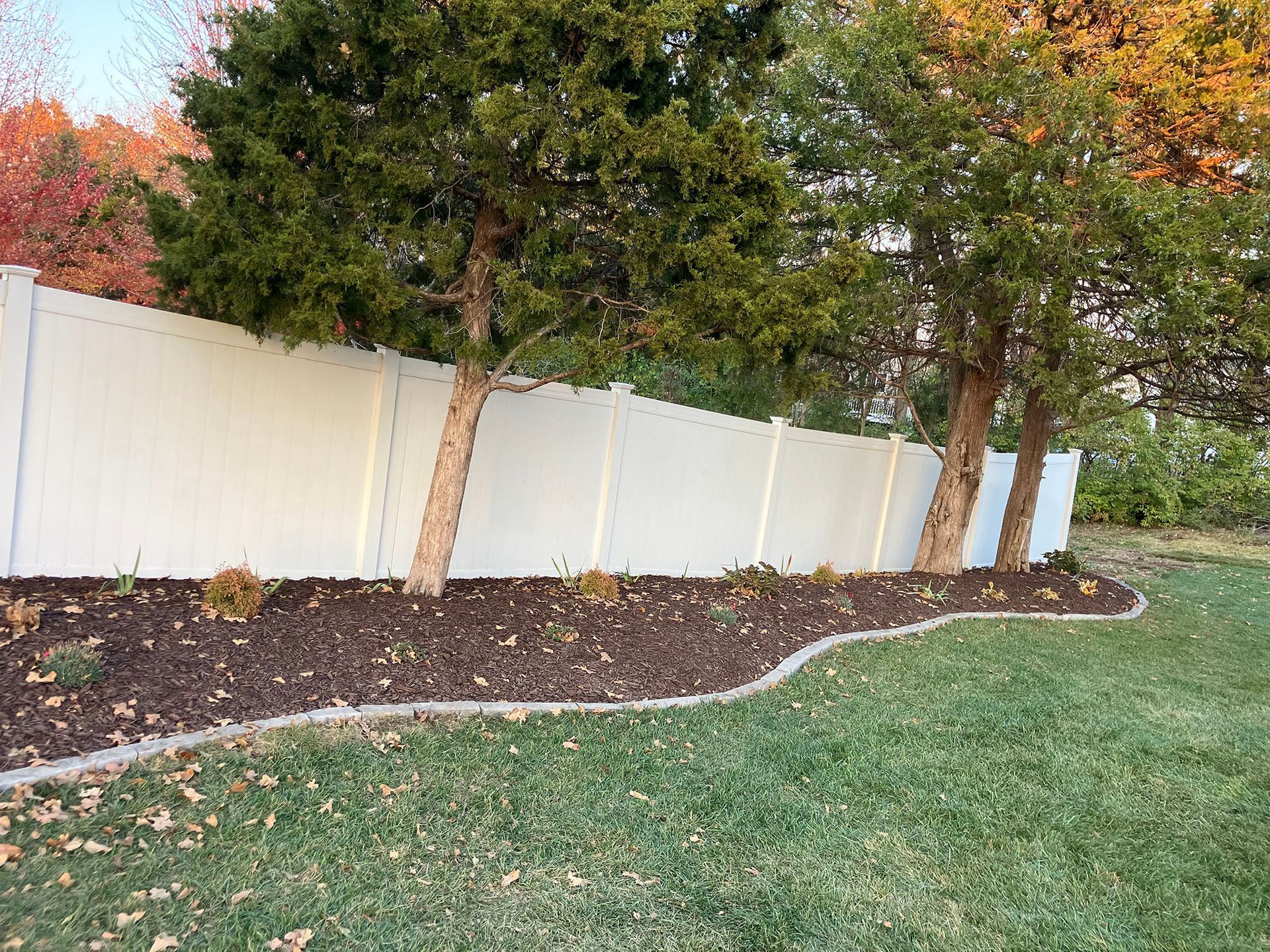 a white fence surrounds a lush green yard with trees and mulch