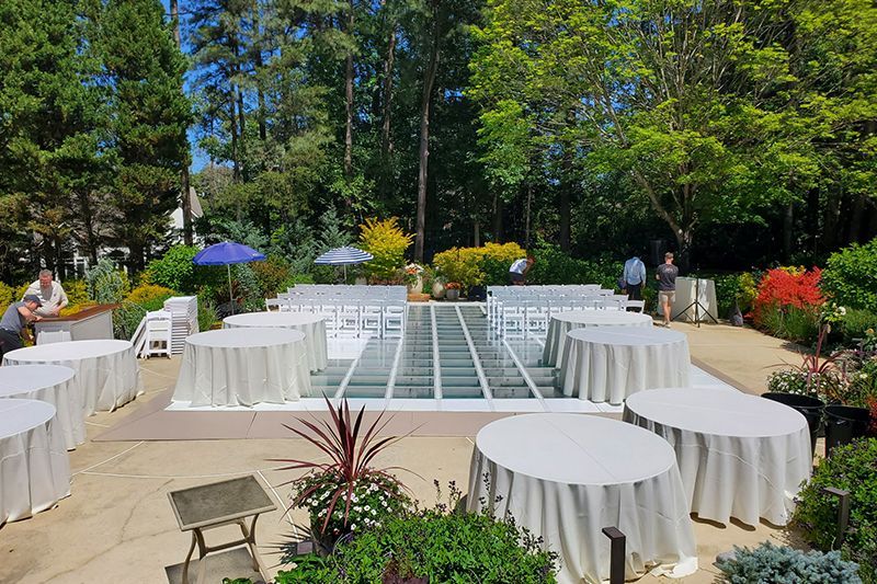 Outdoor garden reception with round white tables and chairs around a glass dance floor, surrounded by trees and flowers