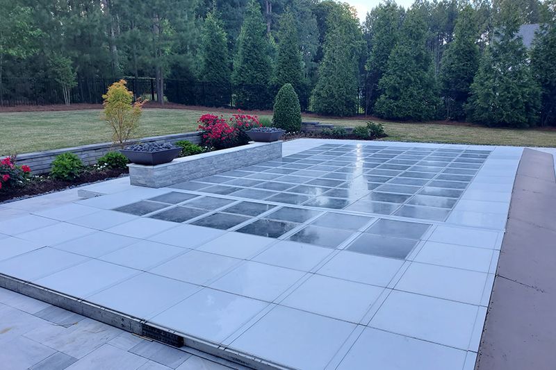 Patio with large glass-tile section, bordered by gray pavers and flower beds beside a grassy yard and trees