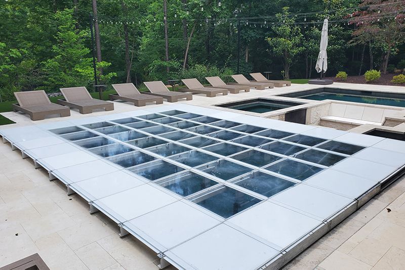 Square outdoor pool covered with white and blue glass panels, surrounded by lounge chairs and greenery