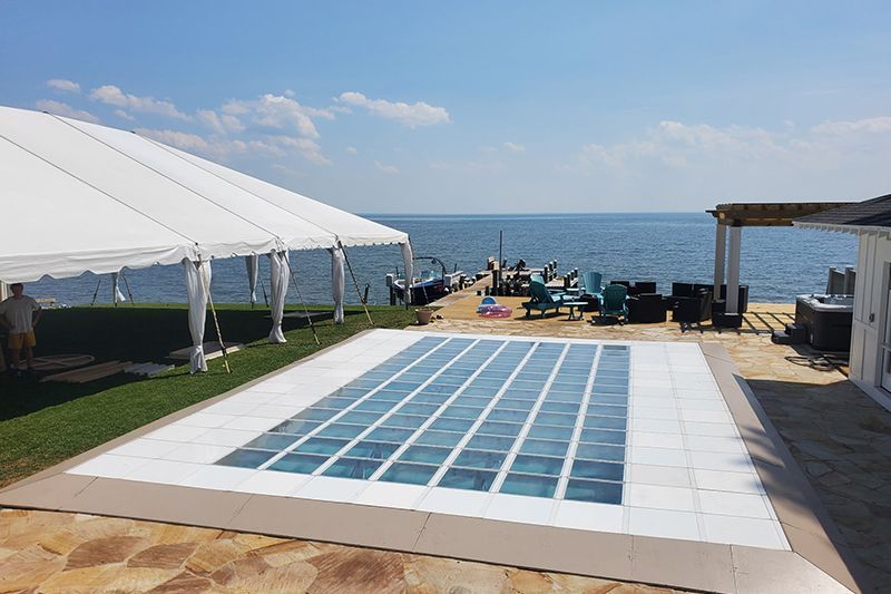 Blue-tiled pool beside a seaside deck with white canopy tents and lounge chairs under a sunny sky