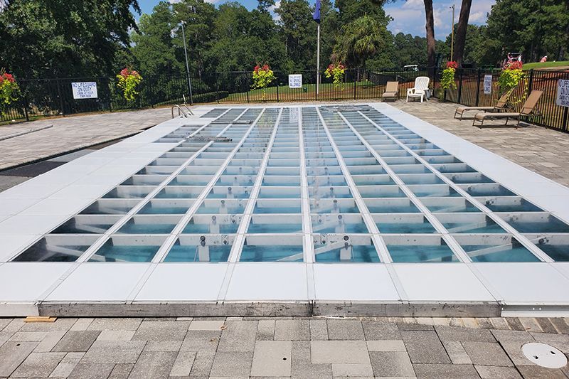 Glass-covered rectangular pool or skylight set in a paved outdoor plaza with trees in the background