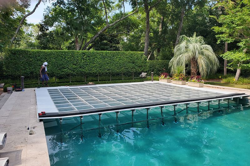 Backyard pool with a closed glass safety cover, surrounded by lush trees and hedges.