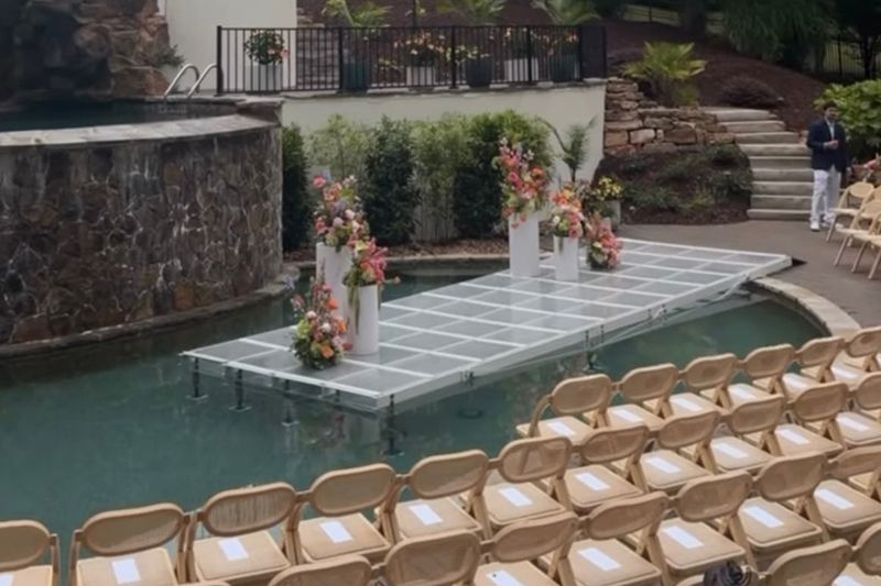 Outdoor wedding setup with floral arch on a floating platform beside rows of tan chairs and a pool.