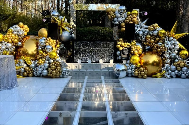 Decorated stage with gold and silver balloon clusters, mirrored tiles, and a reflective walkway outdoors