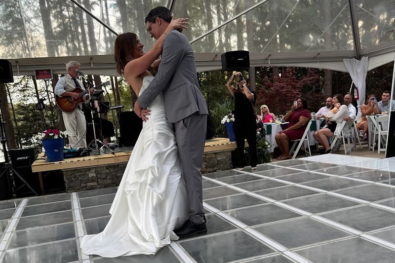 Bride and groom dancing on a clear-tented outdoor reception floor beside a small stage
