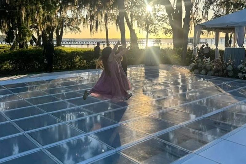 Person in a pink dress on a clear glass floor at sunset, with trees and a lit tent in the background