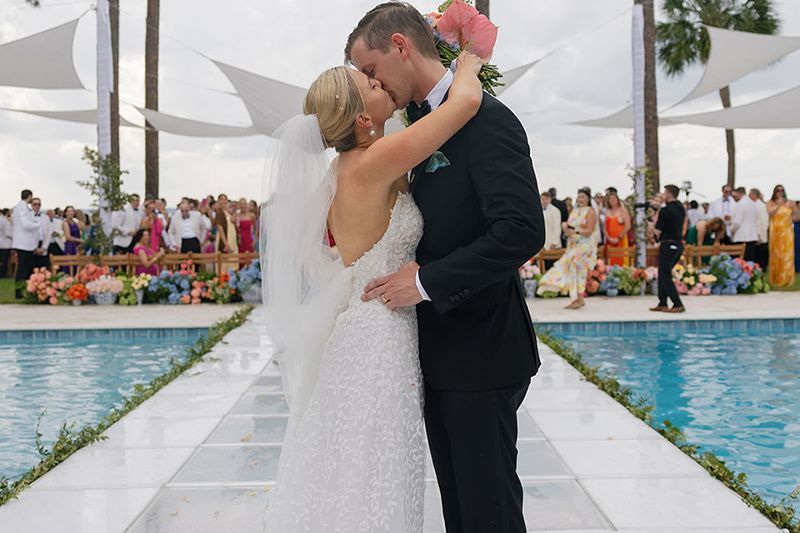 Bride and groom embrace at an outdoor wedding reception beside a pool and floral aisle