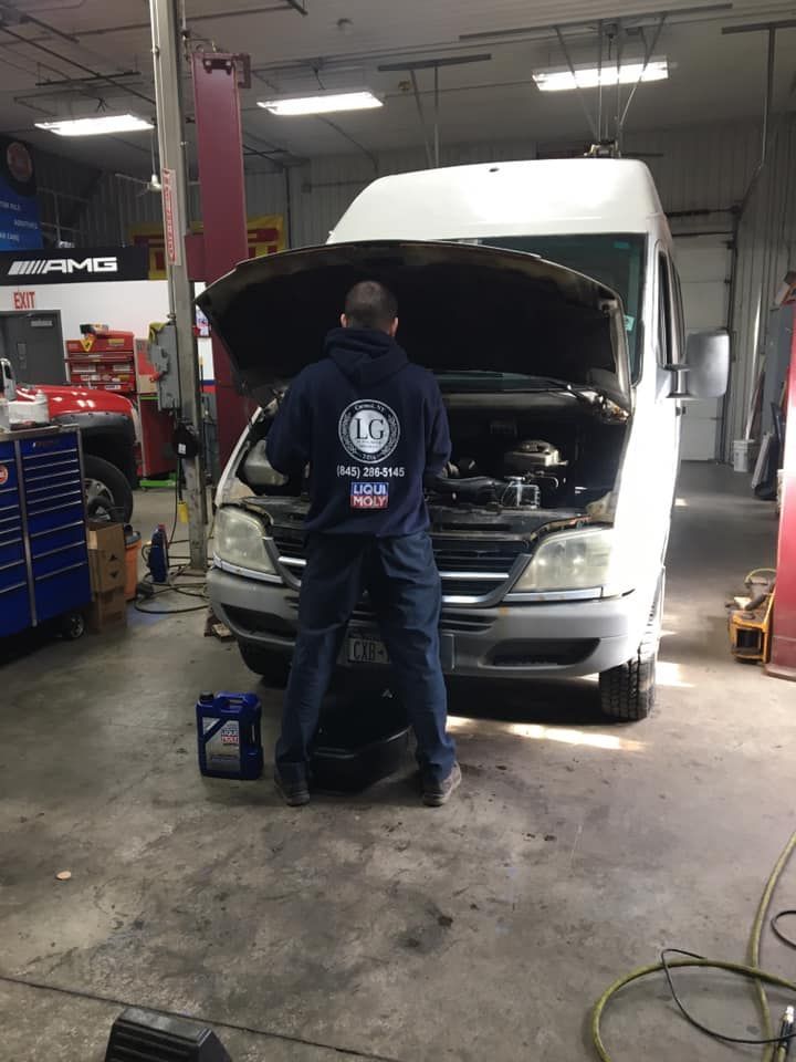 Mechanic working on a van with the hood open in a garage; a bottle of oil is on the ground.
