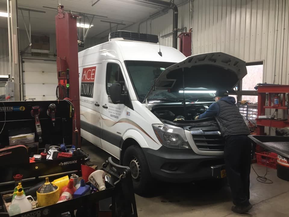 A mechanic working on a white ACE van inside a repair shop. The hood is open.