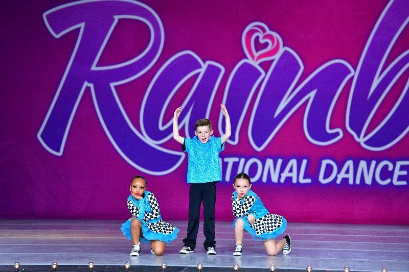 Three young dancers on stage, boy with arms raised, girls kneeling; purple backdrop with